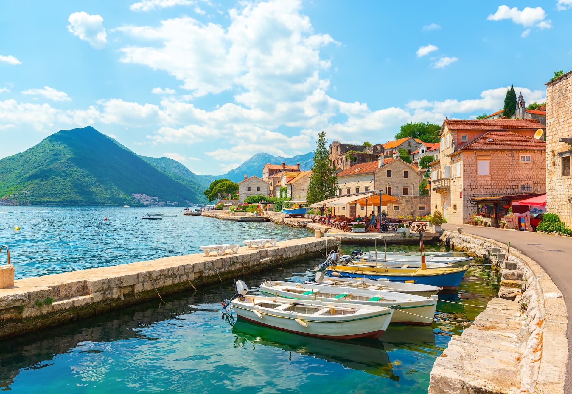 Small boats moored by stone quay in Perast, Montenegro, with stone houses and green mountains.