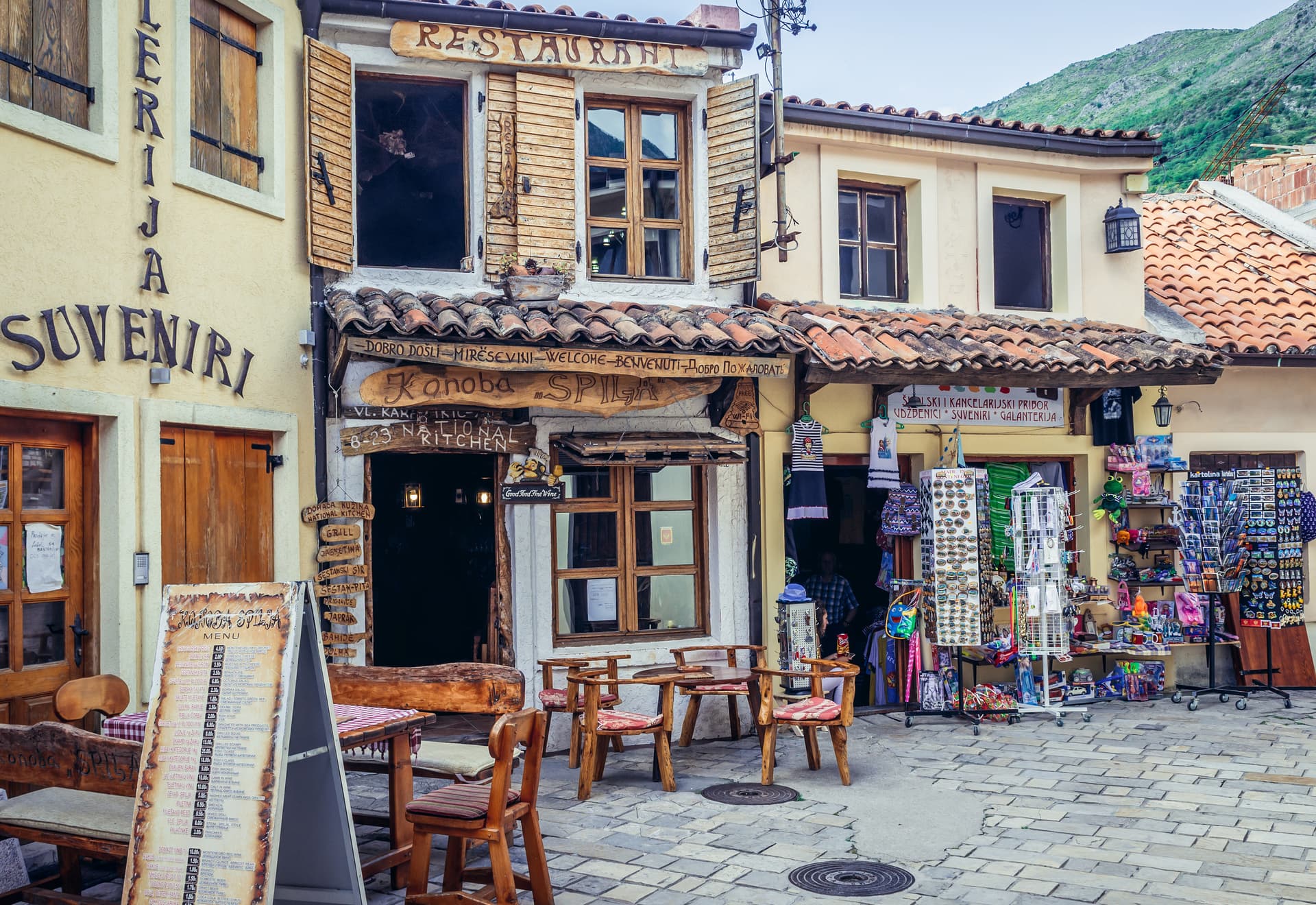 Restaurant and souvenir shops on cobblestone street in Bar, Montenegro, with green mountains visible.