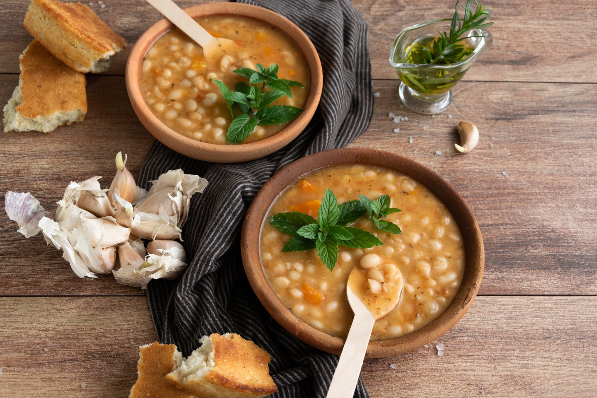 Traditional hot bean soup with cornbread, garlic, and olive oil on a wooden table.