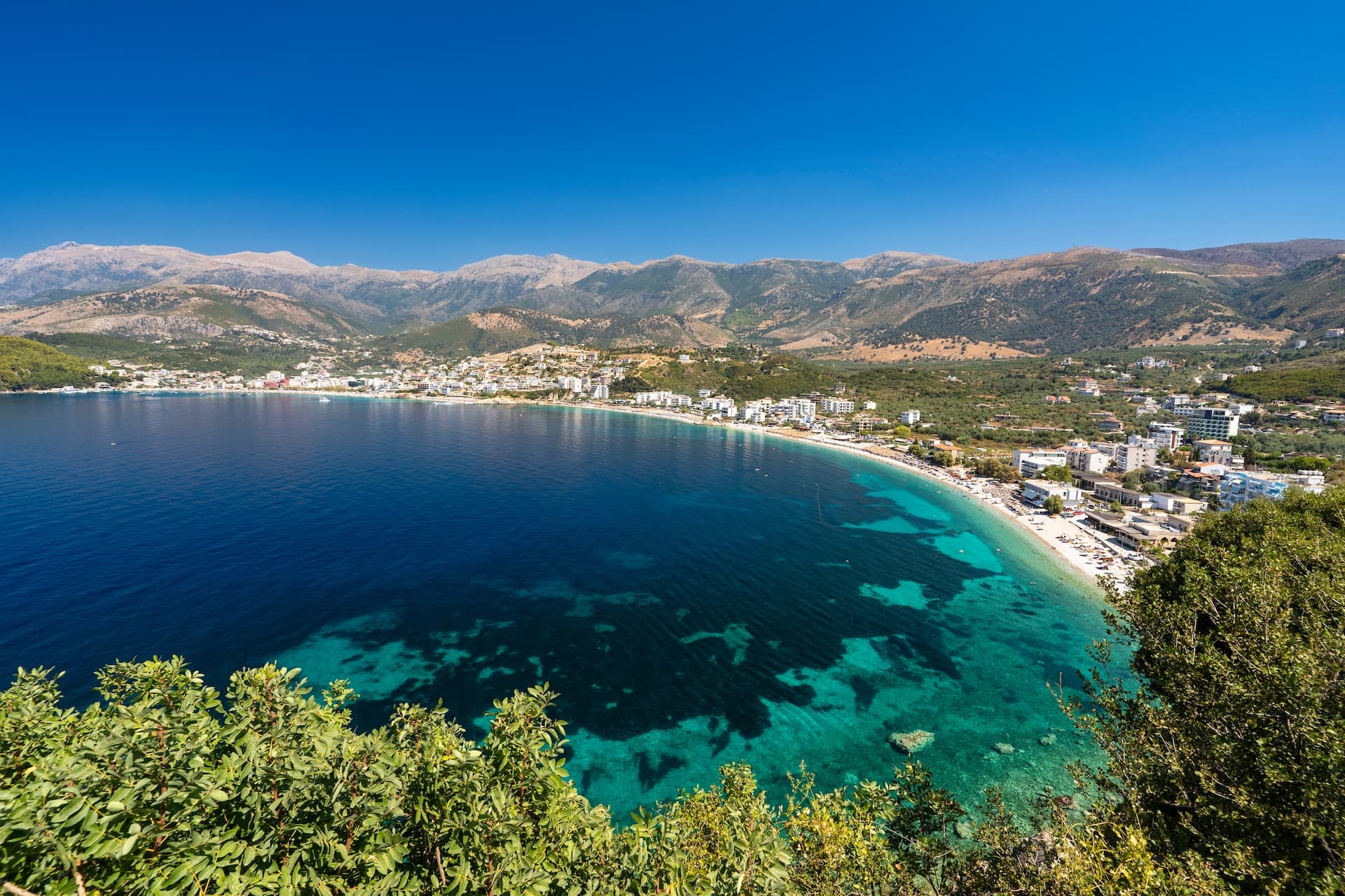 Coastal town of Himare, Albania, with turquoise bay and mountains under a clear blue sky.