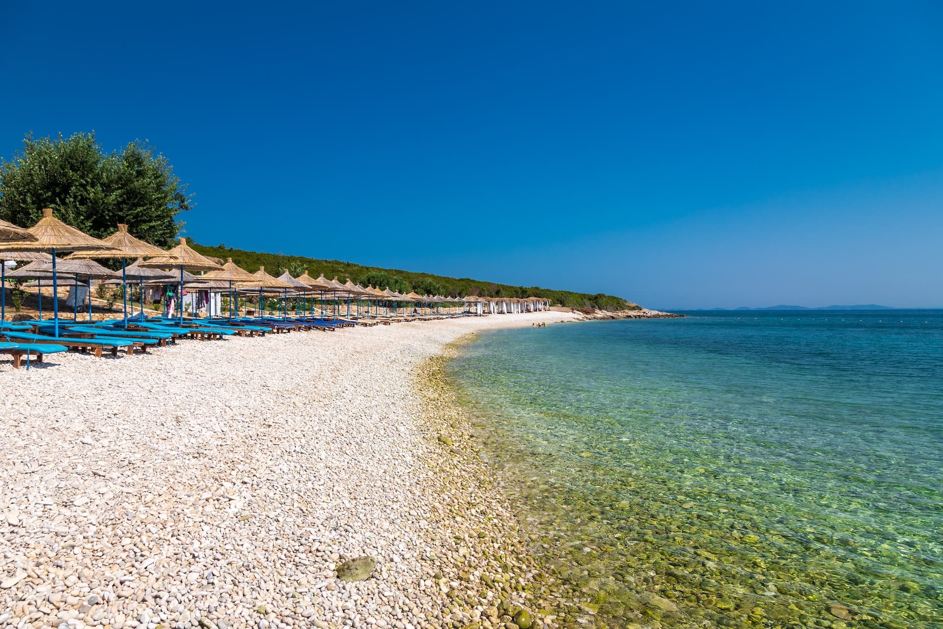 Pebble beach with straw umbrellas and blue loungers on Karaburun Peninsula, Vlore, Albania.