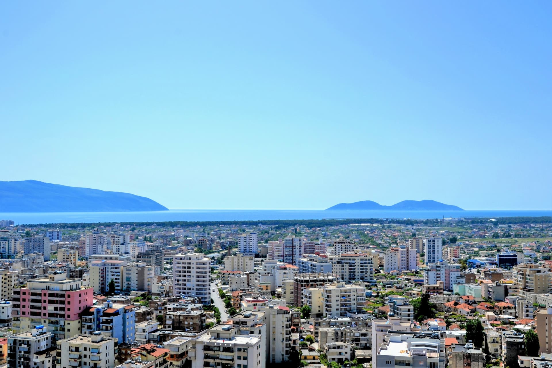 Cityscape of Vlore, Albania, overlooking dense buildings, the sea, and distant blue mountains under a clear sky.