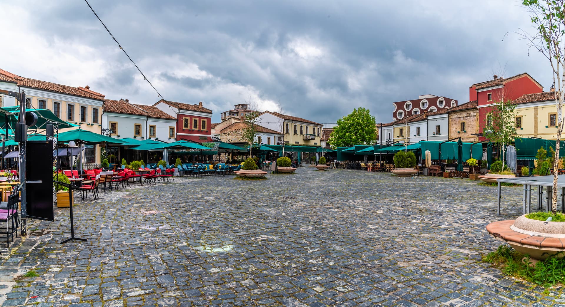 Cobblestone square with outdoor cafe seating under green umbrellas in Korce, Albania.