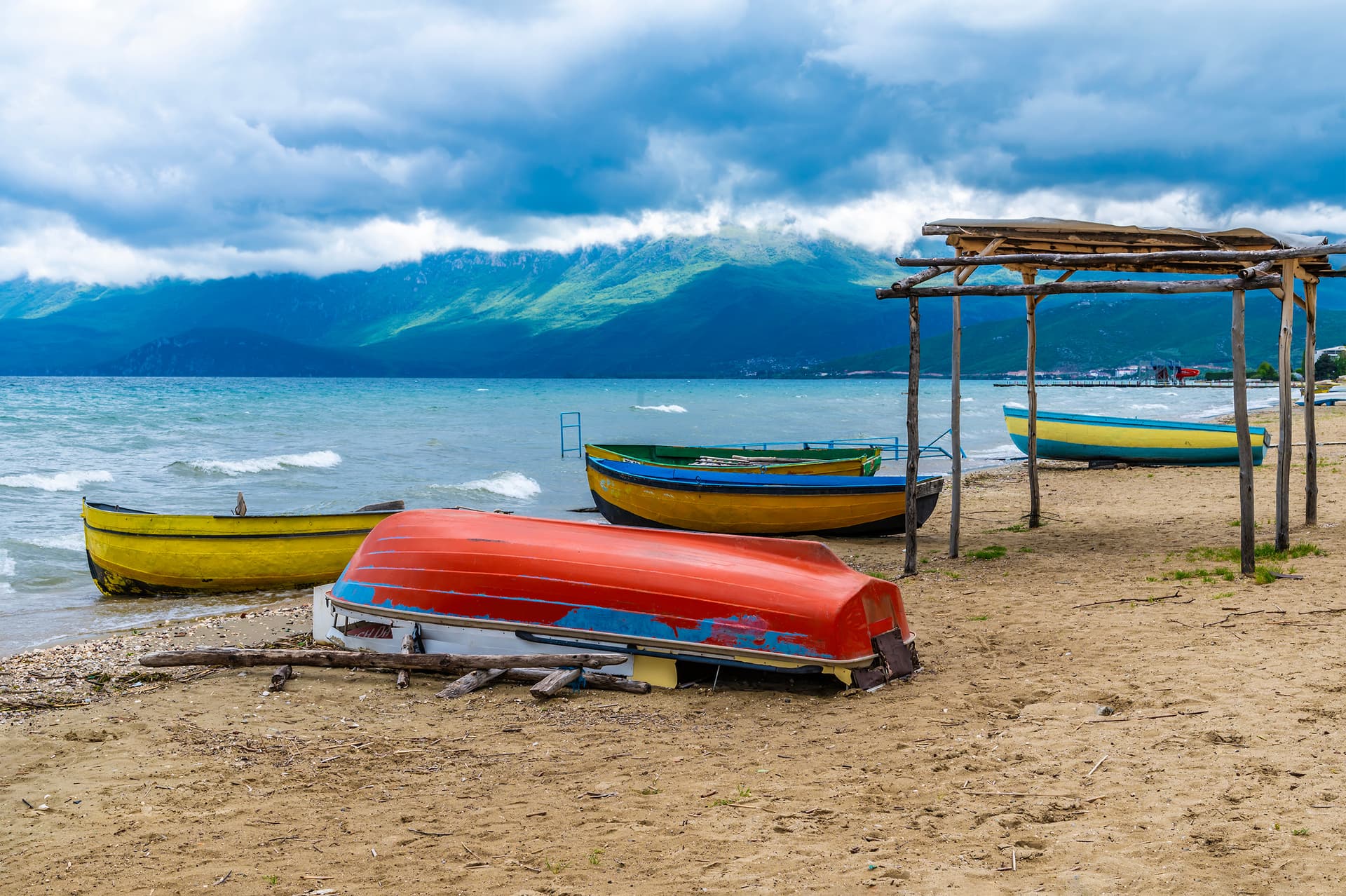 Colorful boats on sandy shore of Lake Ohrid with mountains under cloudy sky.