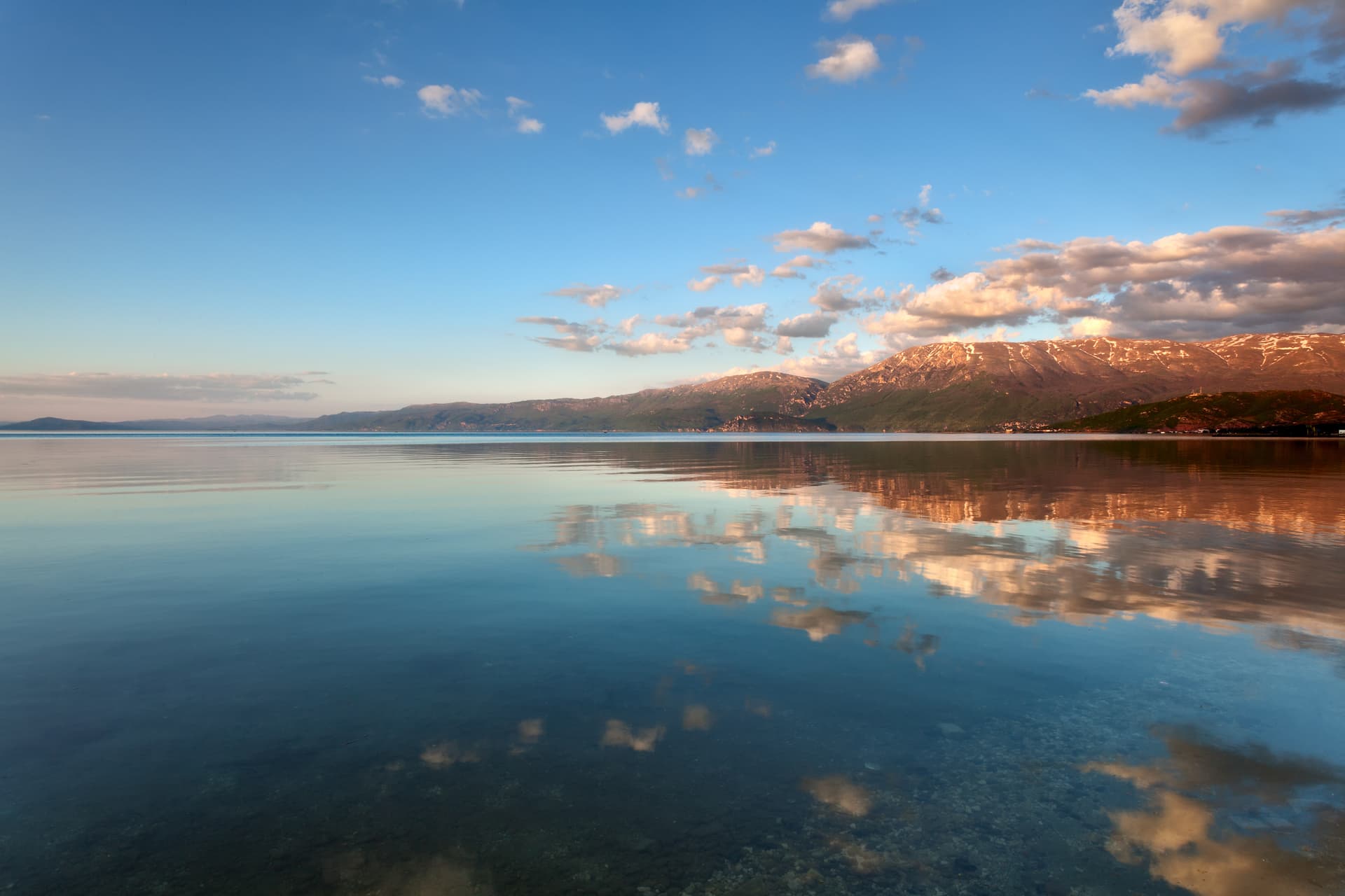 Calm Lake Ohrid, Albania, reflecting mountains and sunset clouds.