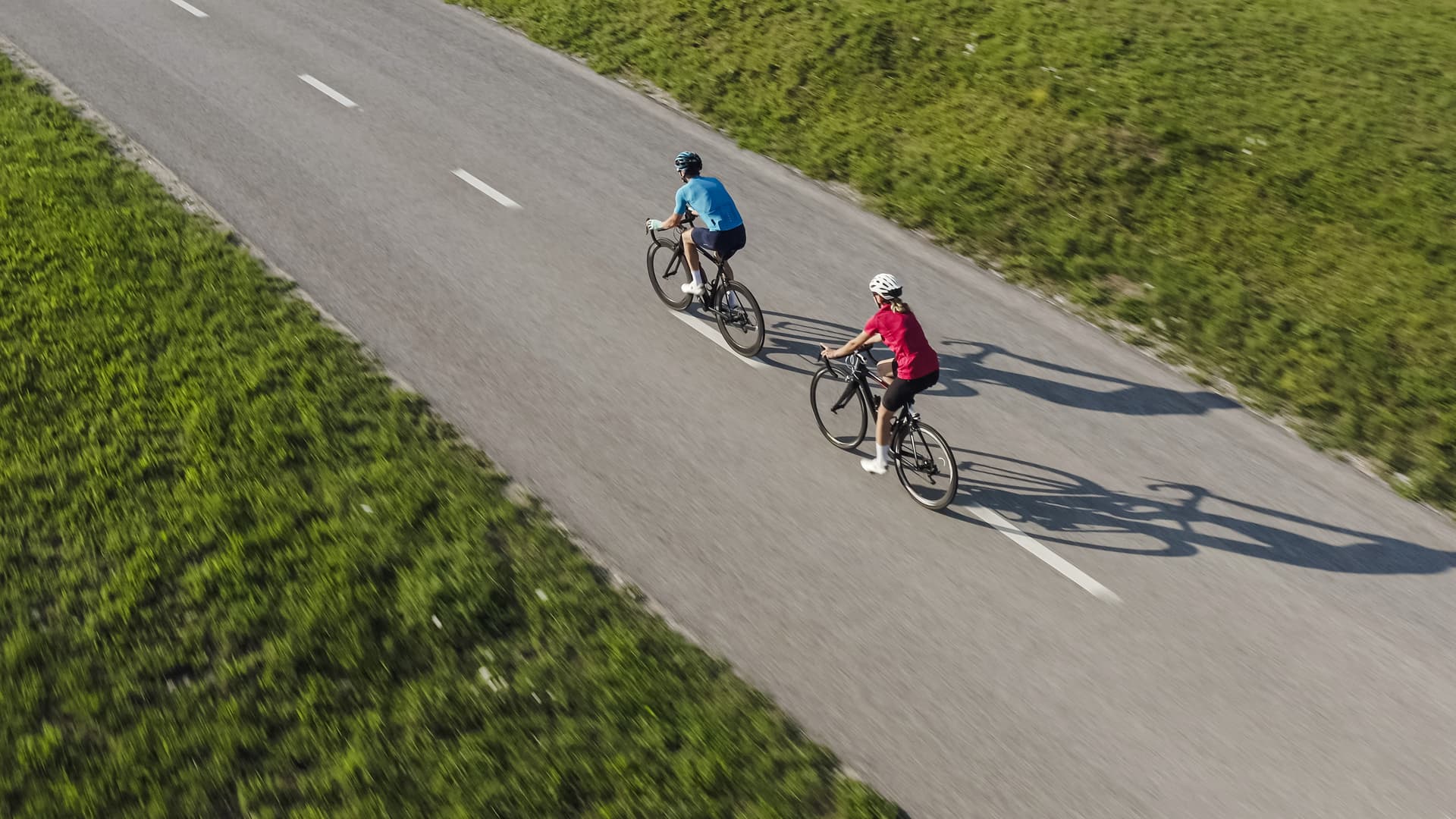 Two cyclists riding road bikes down the middle of a paved road bordered by green grass.