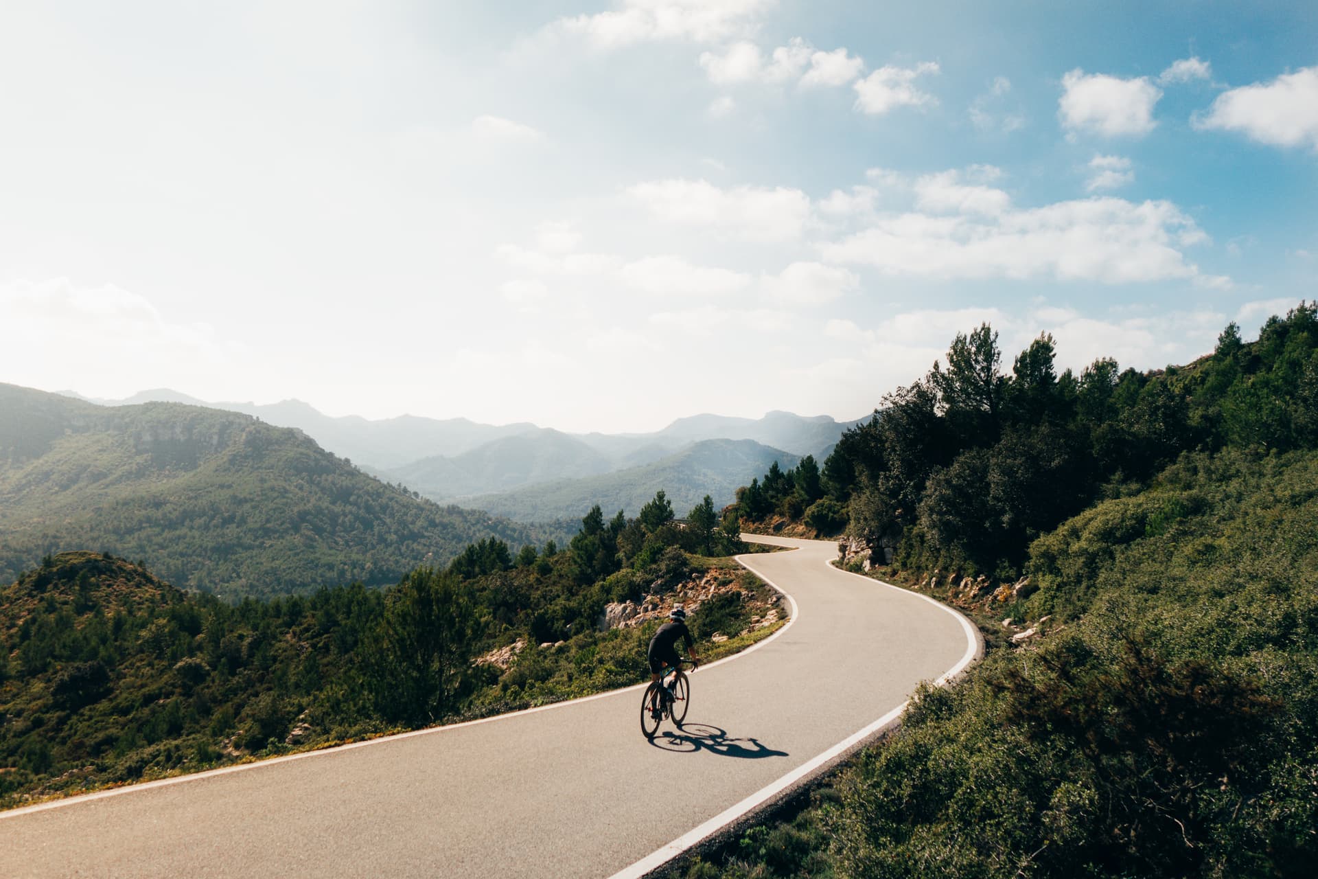 Cyclist descending winding mountain road through green, hazy hills under a bright sky.