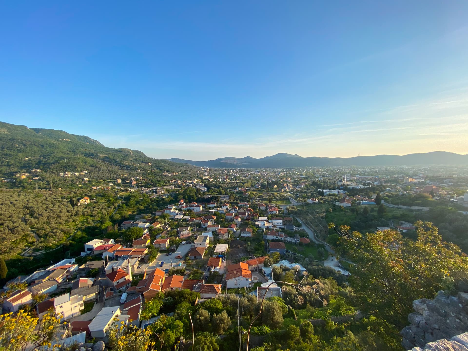 Town nestled in green hillsides under a clear blue sky, with terracotta roofs visible.