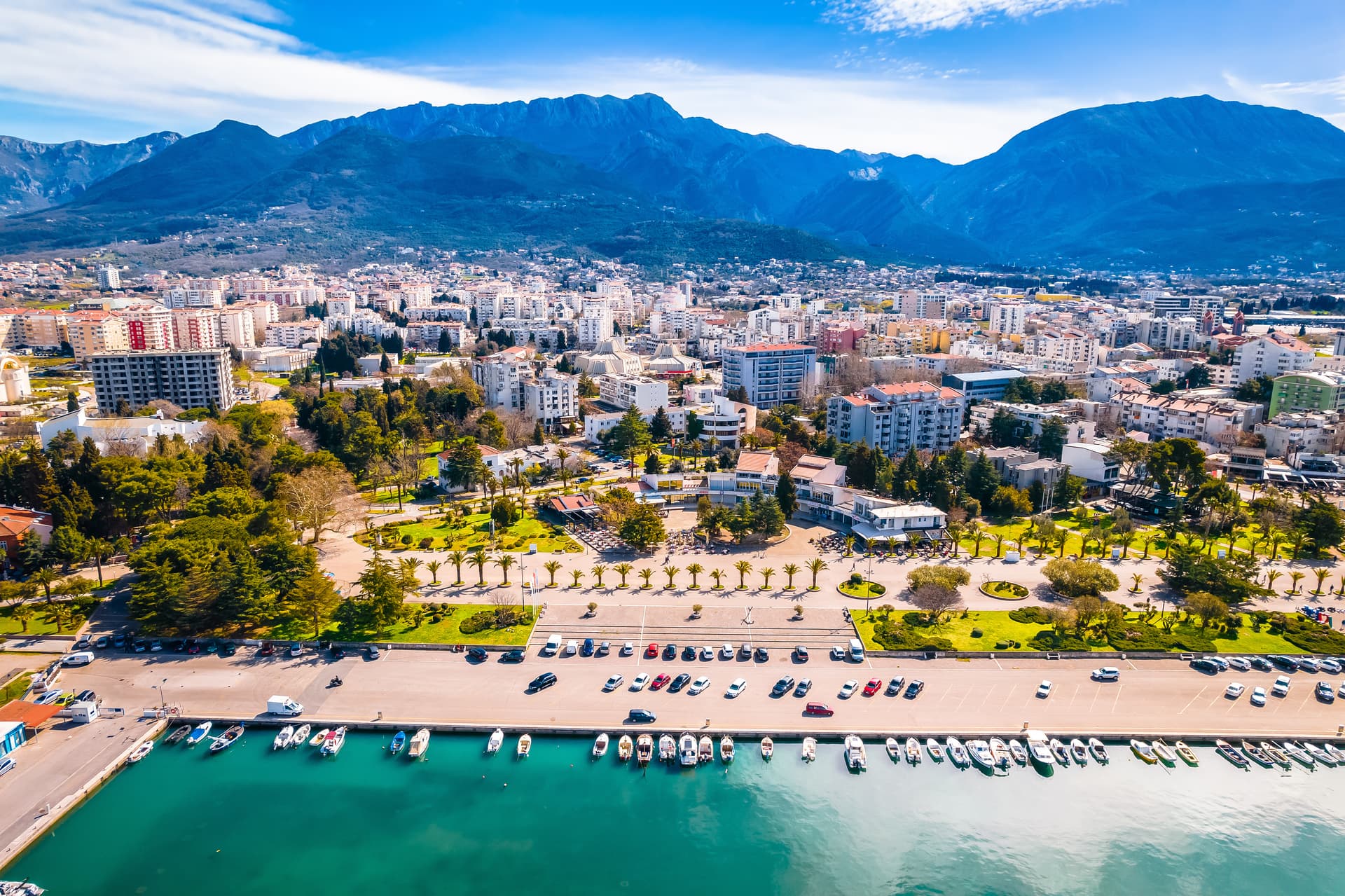 Coastal town with boats docked in turquoise water below large blue mountains in Bar, Montenegro.
