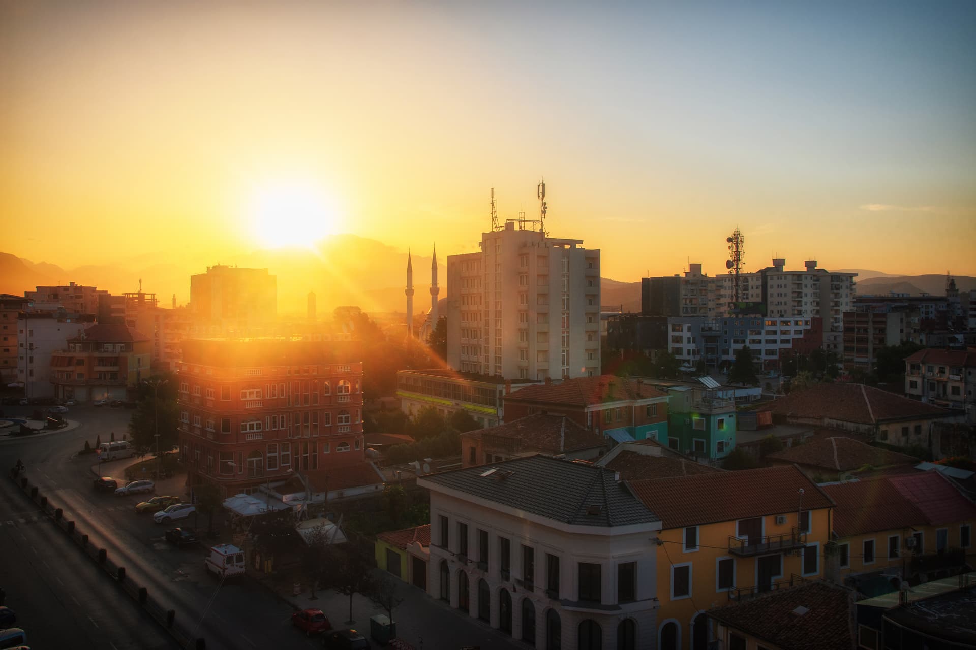 City buildings and mosque minarets silhouetted against a bright sunset in Shkodër, Albania.