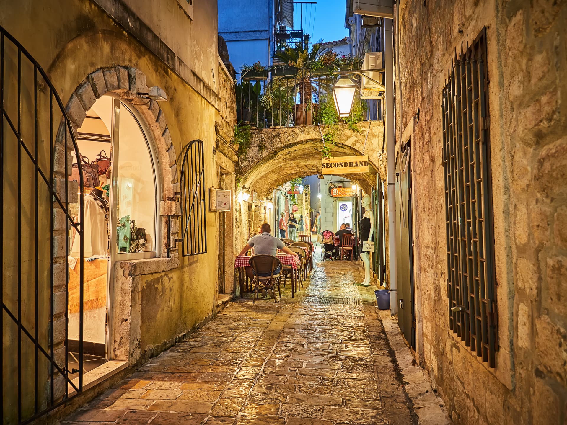 Cobblestone alley in Budva Old Town at night with outdoor dining under an archway.
