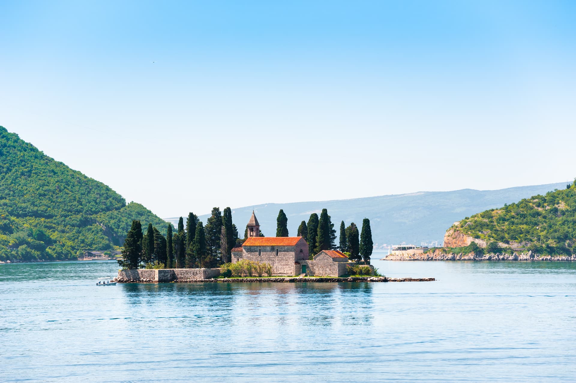 Island church with cypress trees on water, surrounded by green mountains in Perast, Montenegro.