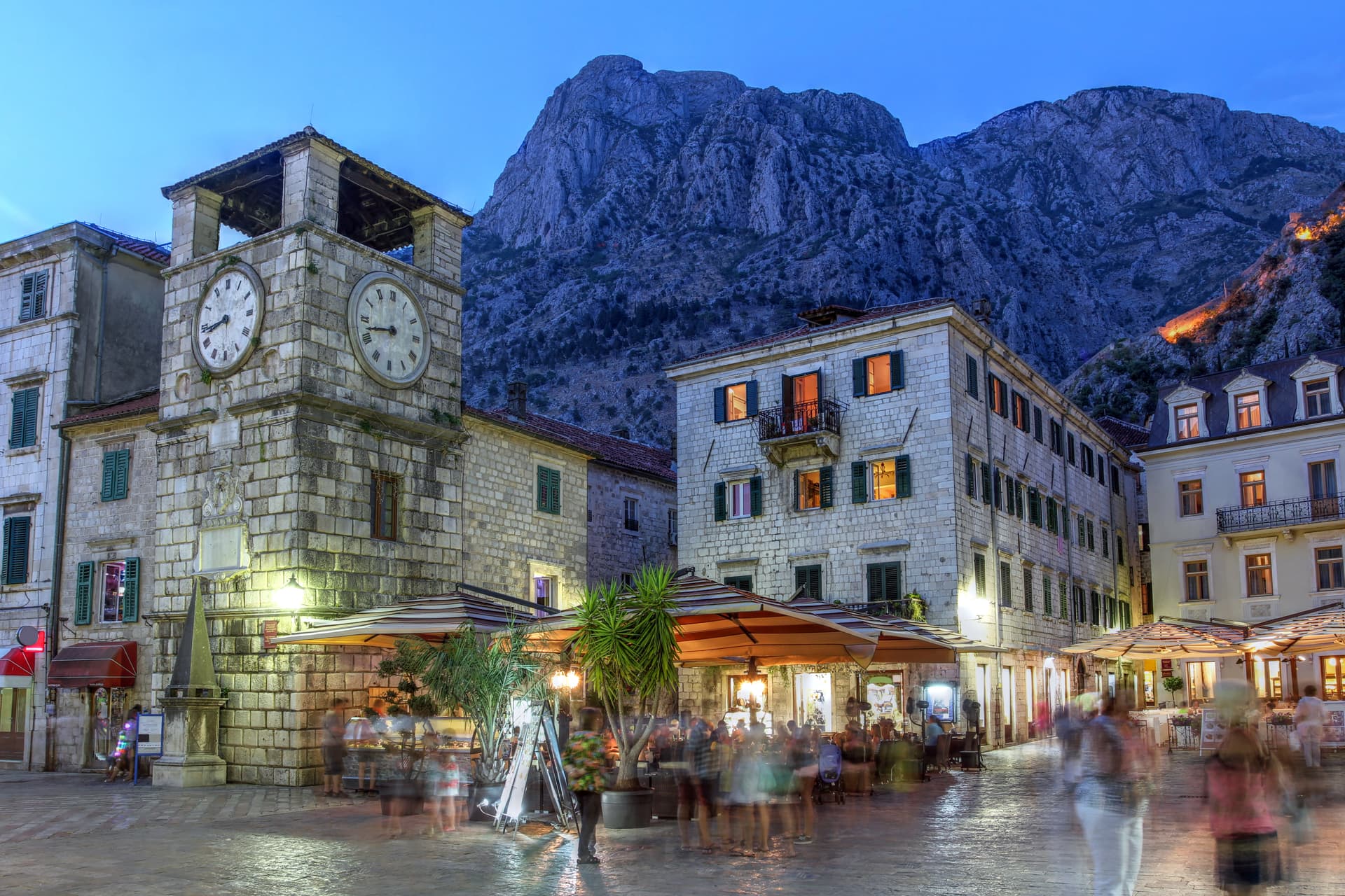 Kotor square at dusk with illuminated clock tower and steep mountain backdrop in Montenegro.